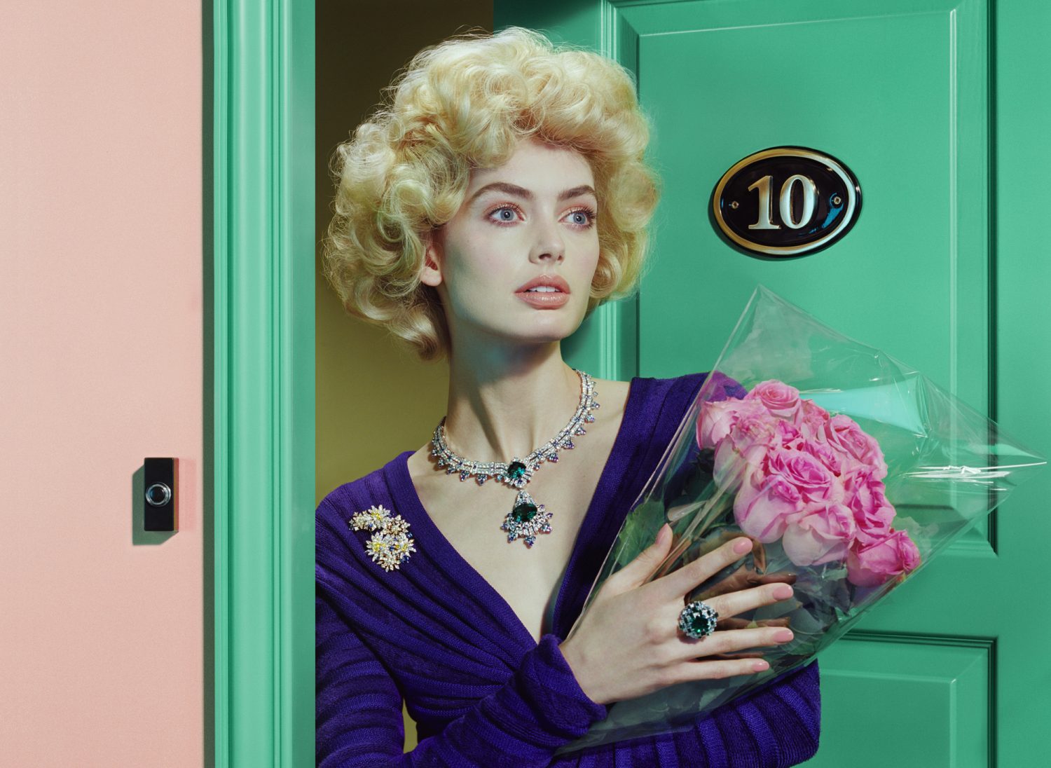 Adult female model posing with pink roses in front of a door and wearing jewelry in a fashion or beauty editorial style photo.