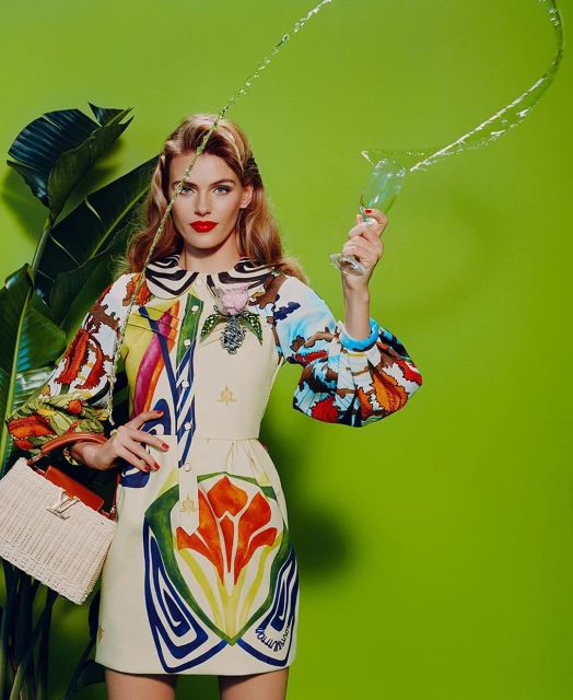 A fair-skinned adult woman with red lipstick poses in a colorful patterned dress, holding a champagne glass with liquid against a green backdrop.