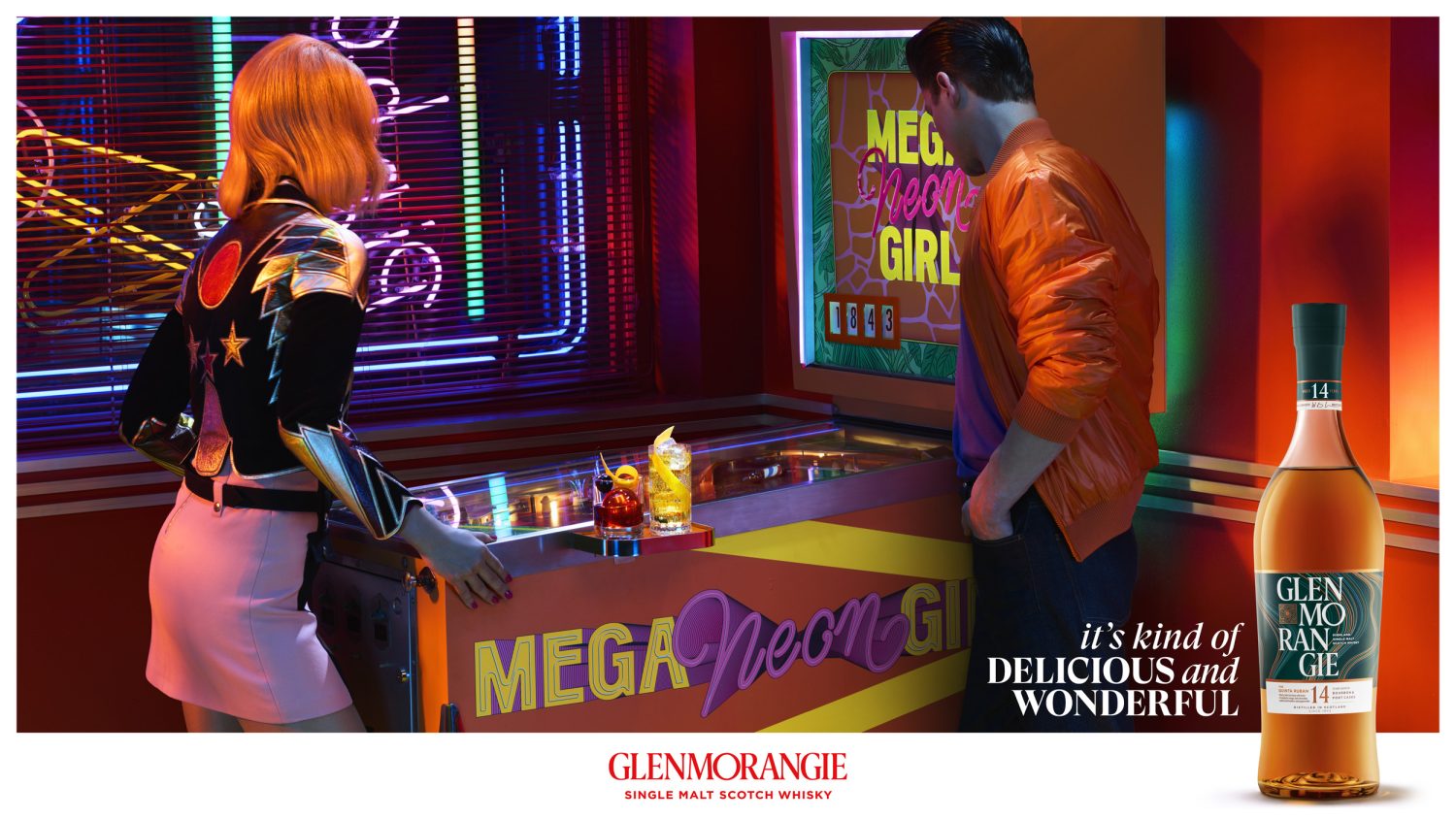 Two people at a Mega Neon Girl pinball machine, a table of cocktails, and a bottle of Glenmorangie Scotch Whisky in a vibrant retro bar setting.