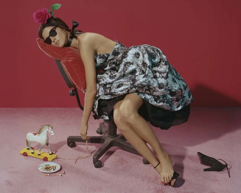 Fashion shot of a woman in a printed dress reclining on an office chair with toys and a red background in a studio setting.