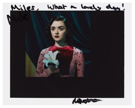 Glamorous portrait of a young woman with red lips, holding a white lily in front of a dark teal and yellow backdrop with handwritten text.