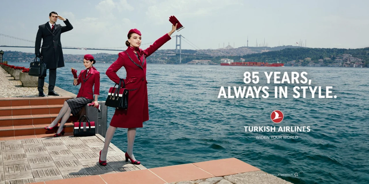 An outdoor advertisement for Turkish Airlines featuring three airline employees dressed in formal uniforms standing by the sea with a bridge.