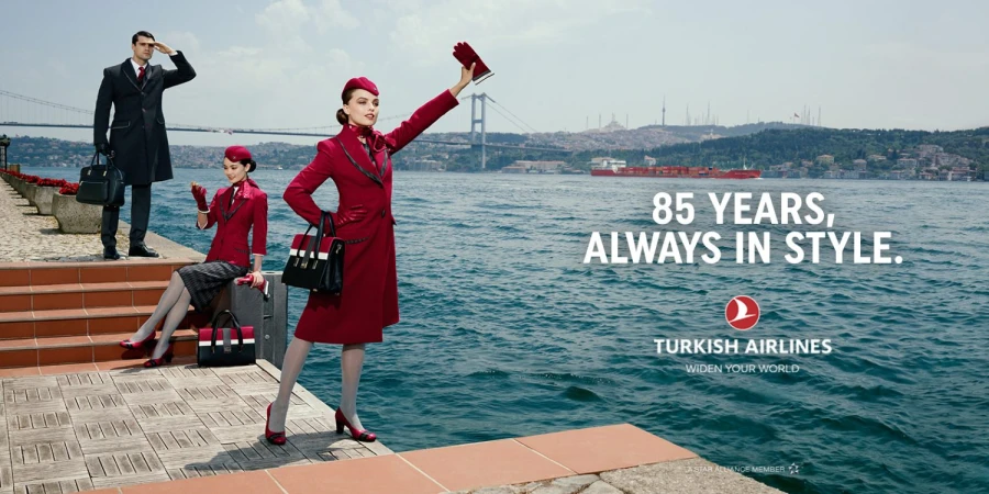 An outdoor advertisement for Turkish Airlines featuring three airline employees dressed in formal uniforms standing by the sea with a bridge.