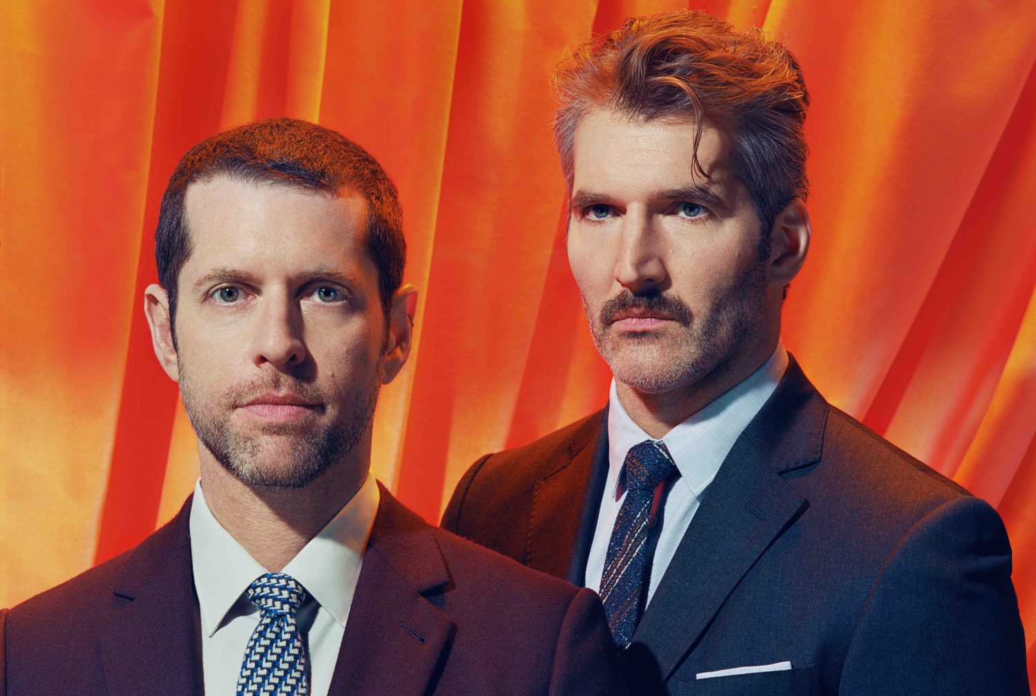 Two adult males, one with a mustache and grey hair, posing in suits against an orange backdrop for a formal studio portrait.