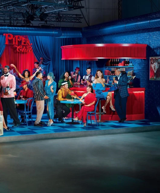 Large group of diverse adults dressed in 1920s attire inside a red and blue photo studio set with tables and chairs, plus 'Piper Club' signage.