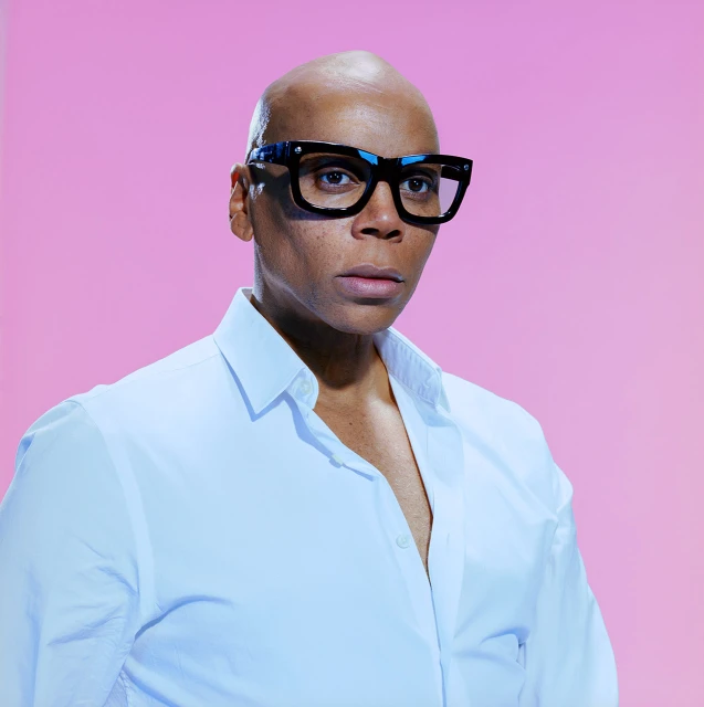 RuPaul in a studio portrait, wearing a white button-down shirt and black square-framed glasses, against a gradient pink background.