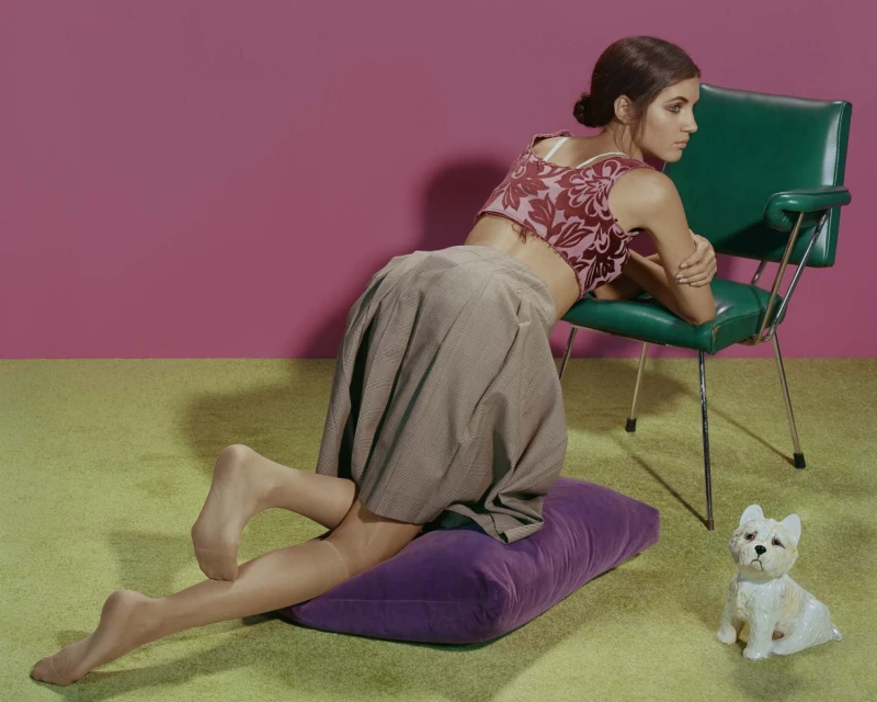 Brunette woman kneels on a purple pillow next to a green chair and dog figurine, posing in a studio setting with a pink backdrop.