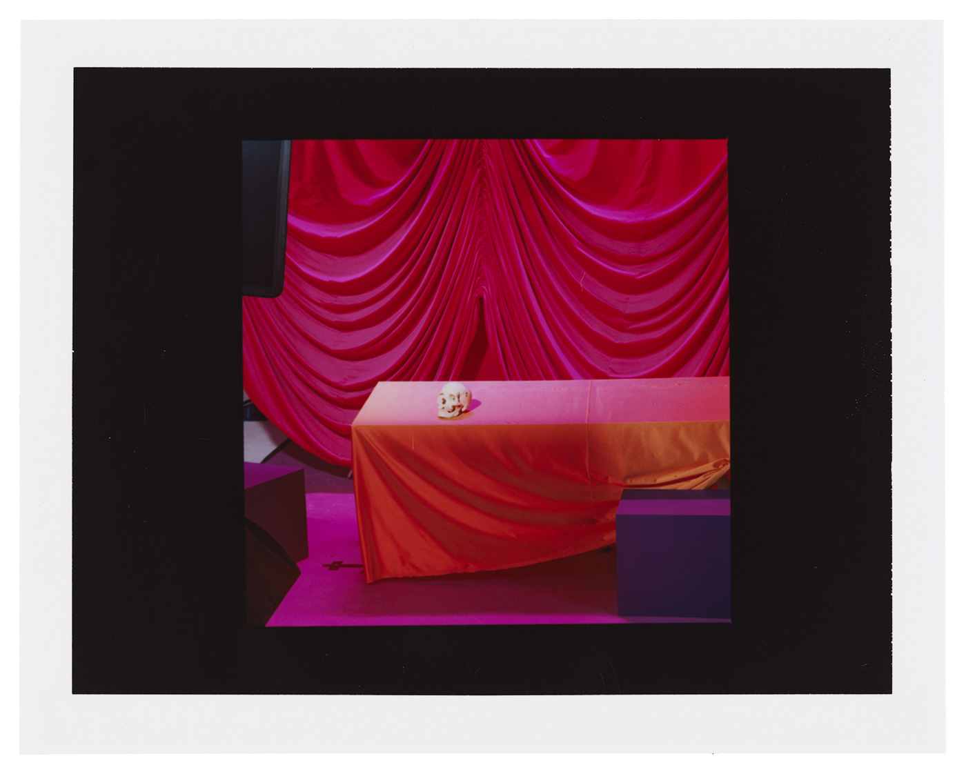 A table covered with an orange and pink cloth holds an object, set against a backdrop of draped magenta fabric indoors.