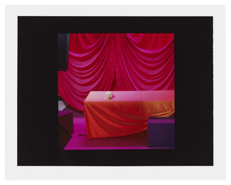 A table covered with an orange and pink cloth holds an object, set against a backdrop of draped magenta fabric indoors.