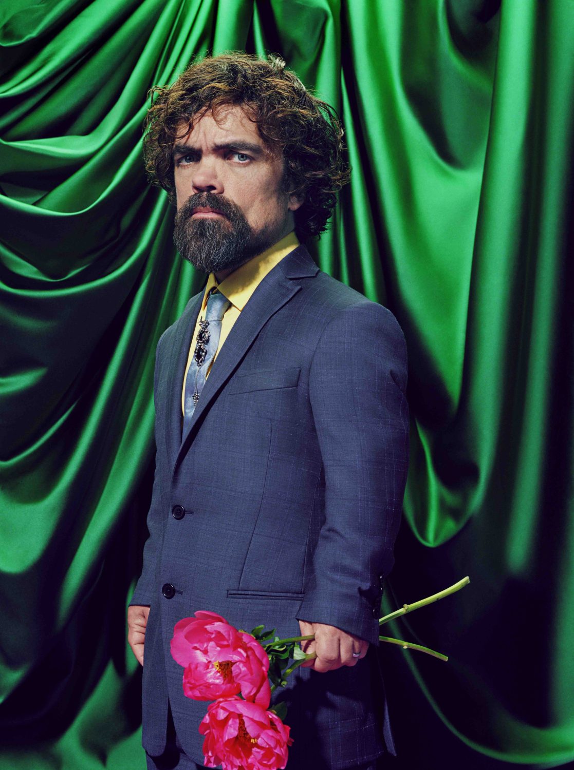 Studio portrait of a man in a blue suit holding two pink peonies against draped green fabric, featuring a formal and elegant aesthetic.