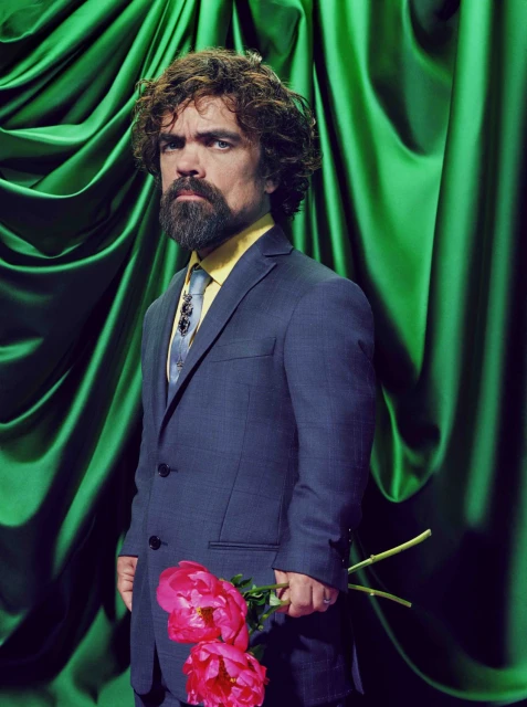 Studio portrait of a man in a blue suit holding two pink peonies against draped green fabric, featuring a formal and elegant aesthetic.