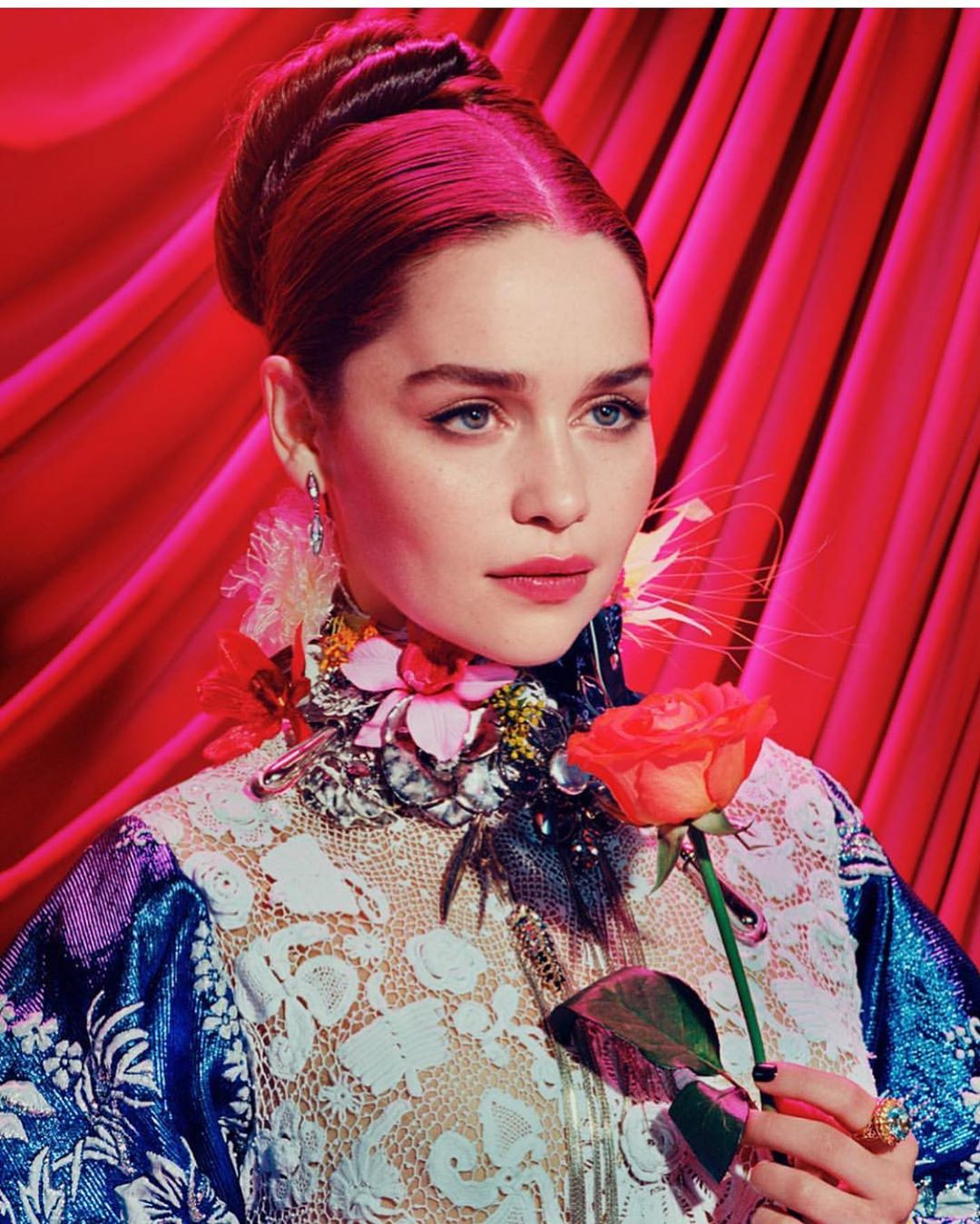 Portrait of a red-haired adult woman with rose wearing a lace top against a red draped background in a studio setting.
