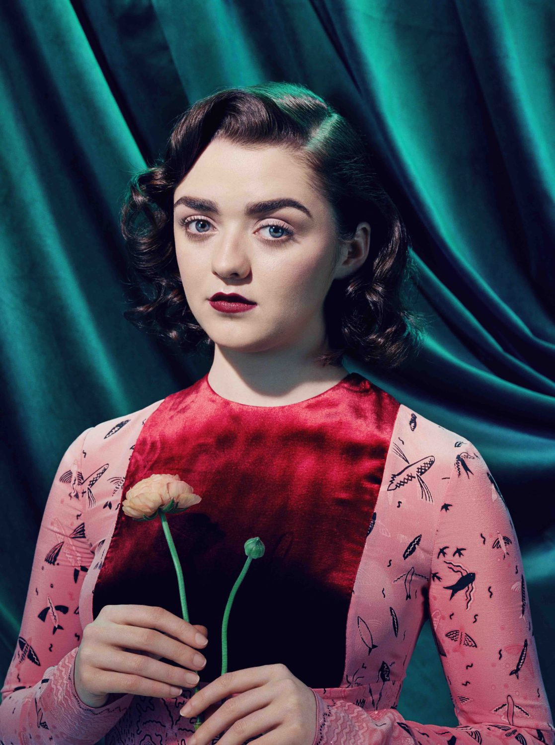 Portrait of a young woman with dark hair, wearing a pink dress, and holding a flower against a teal fabric background in a studio.
