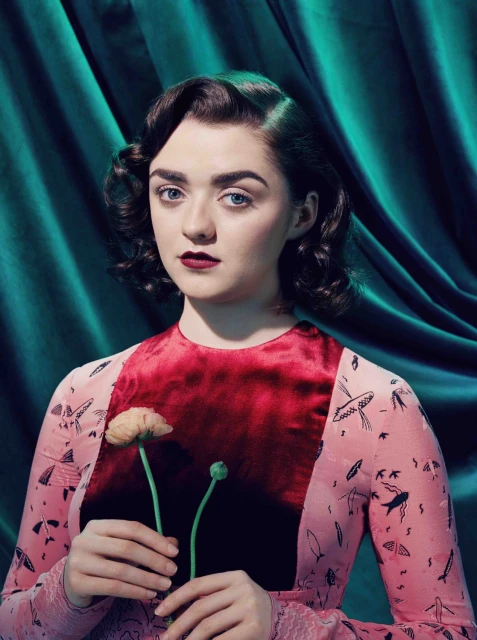 Portrait of a young woman with dark hair, wearing a pink dress, and holding a flower against a teal fabric background in a studio.