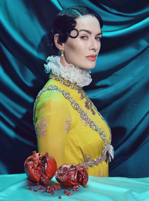 Elegant studio portrait of a woman in a yellow dress with a white ruffled collar, adorned with jewels, alongside pomegranates.