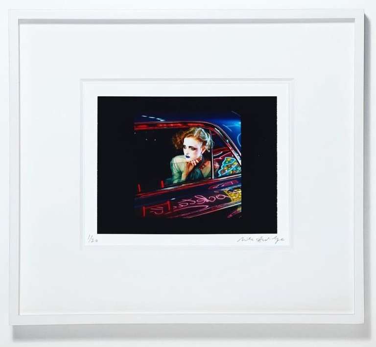 Framed image of a person with dramatic makeup sitting in a car with reflective neon lights, framed with a white matte and frame.