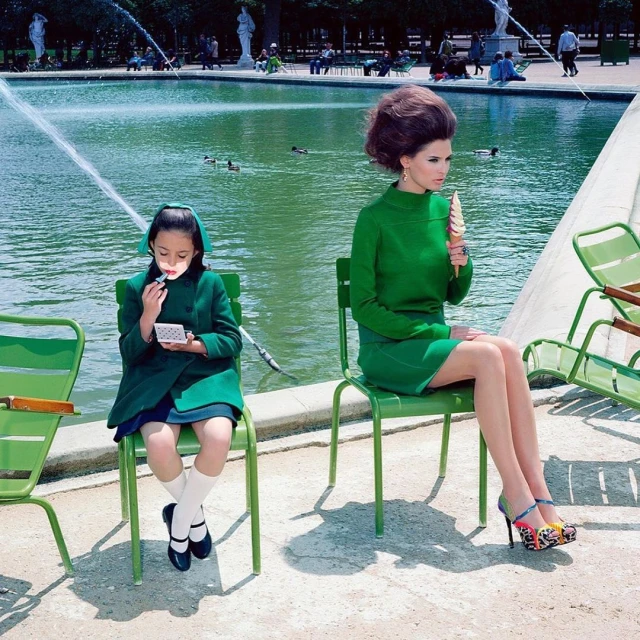 Two models sit in a park setting. One is a child applying makeup while the other is an adult eating an ice cream cone.