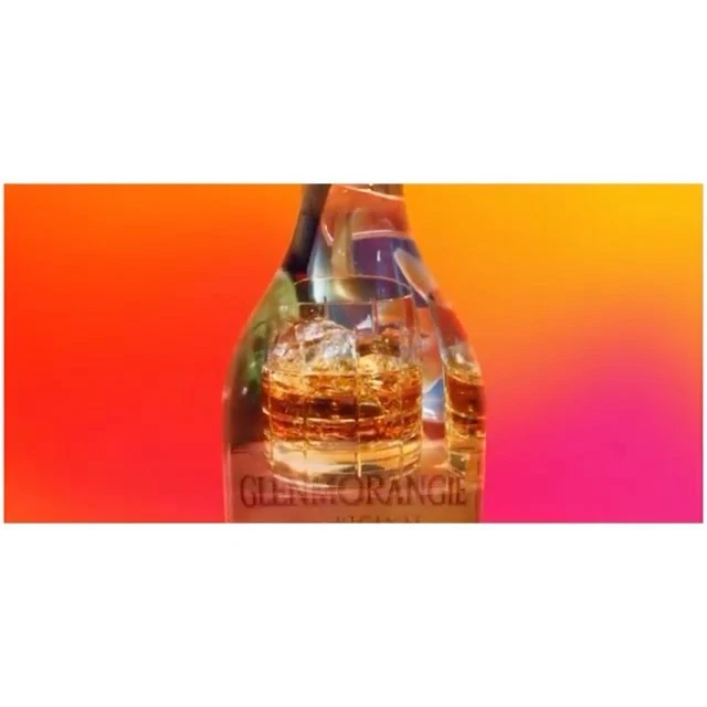 Whiskey bottle and glass on a blurred background featuring gradient tones of orange and pink. The whiskey glass features ice.