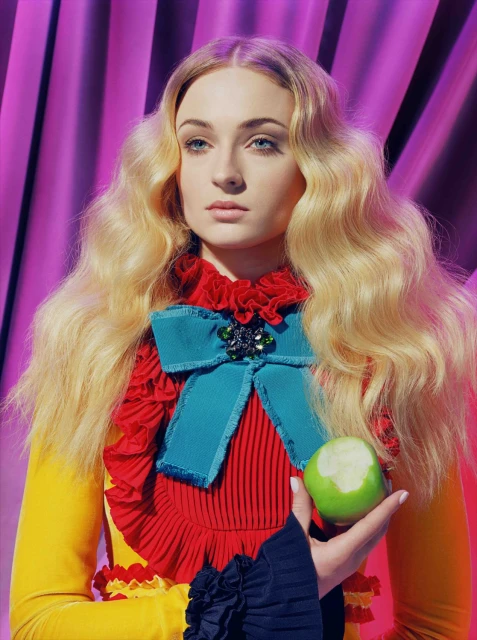 Blonde woman with fair skin poses holding a green apple with a bite taken out of it in front of a pink backdrop.