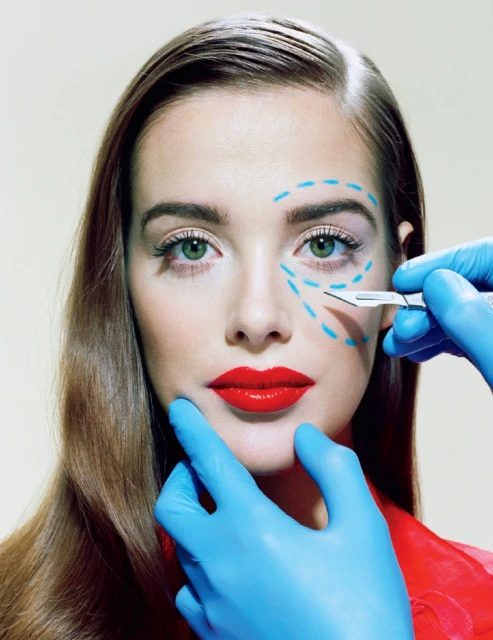 Close-up of a woman's face with blue cosmetic surgery markings, a scalpel held in a blue-gloved hand near her eye. Red lips.