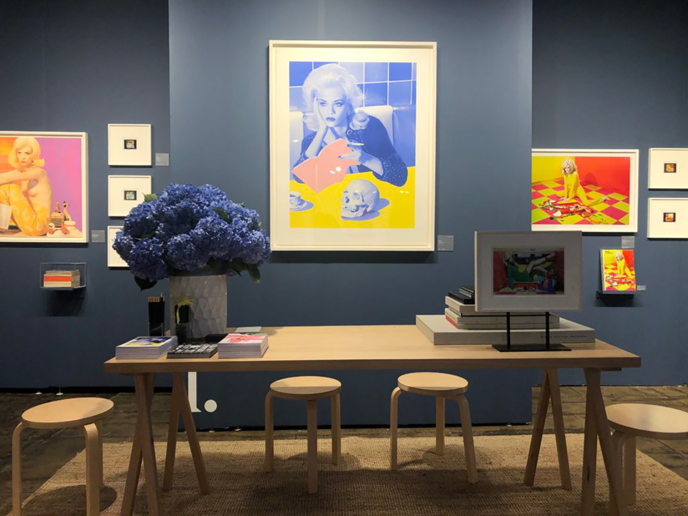 Artwork installation with framed prints of women and still life on a blue wall above a light wood table and stools, gallery exhibition.