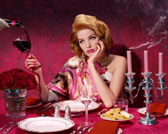 Glamorous woman in pink patterned dress at a dining table with wine glass, roses, silver candelabra, plates and table setting.