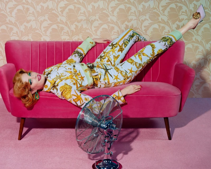 A red-haired woman in patterned pajamas reclines on a pink couch with a vintage fan in a studio setting with floral wallpaper.