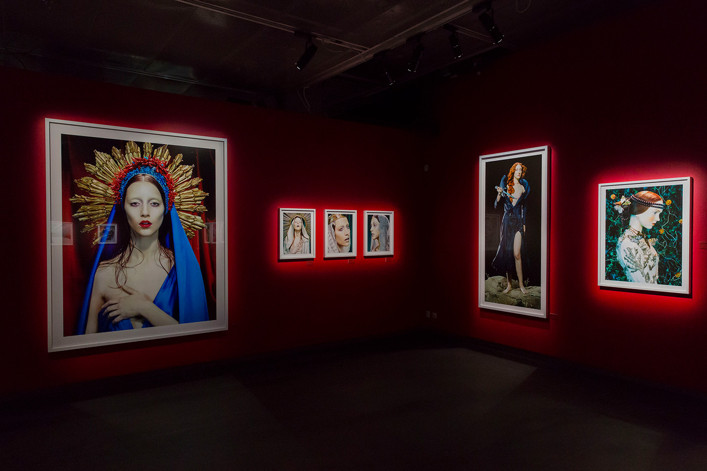 Art gallery featuring portraits of women with red and gold headpieces and blue drapes, displayed on red walls in white frames.