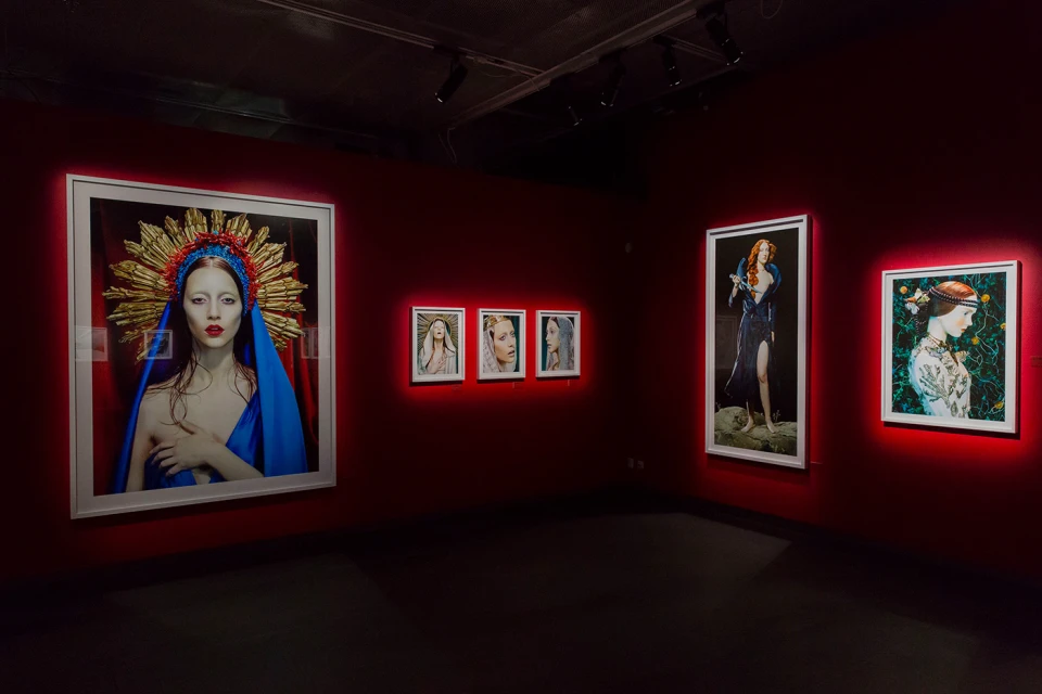 Art gallery featuring portraits of women with red and gold headpieces and blue drapes, displayed on red walls in white frames.