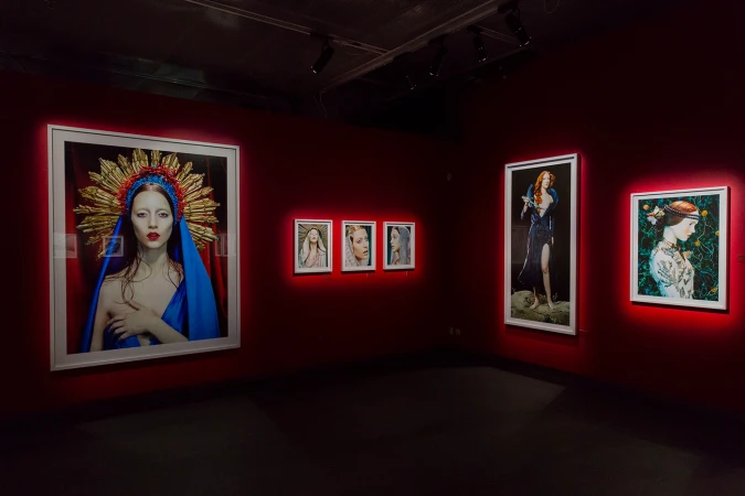Art gallery featuring portraits of women with red and gold headpieces and blue drapes, displayed on red walls in white frames.