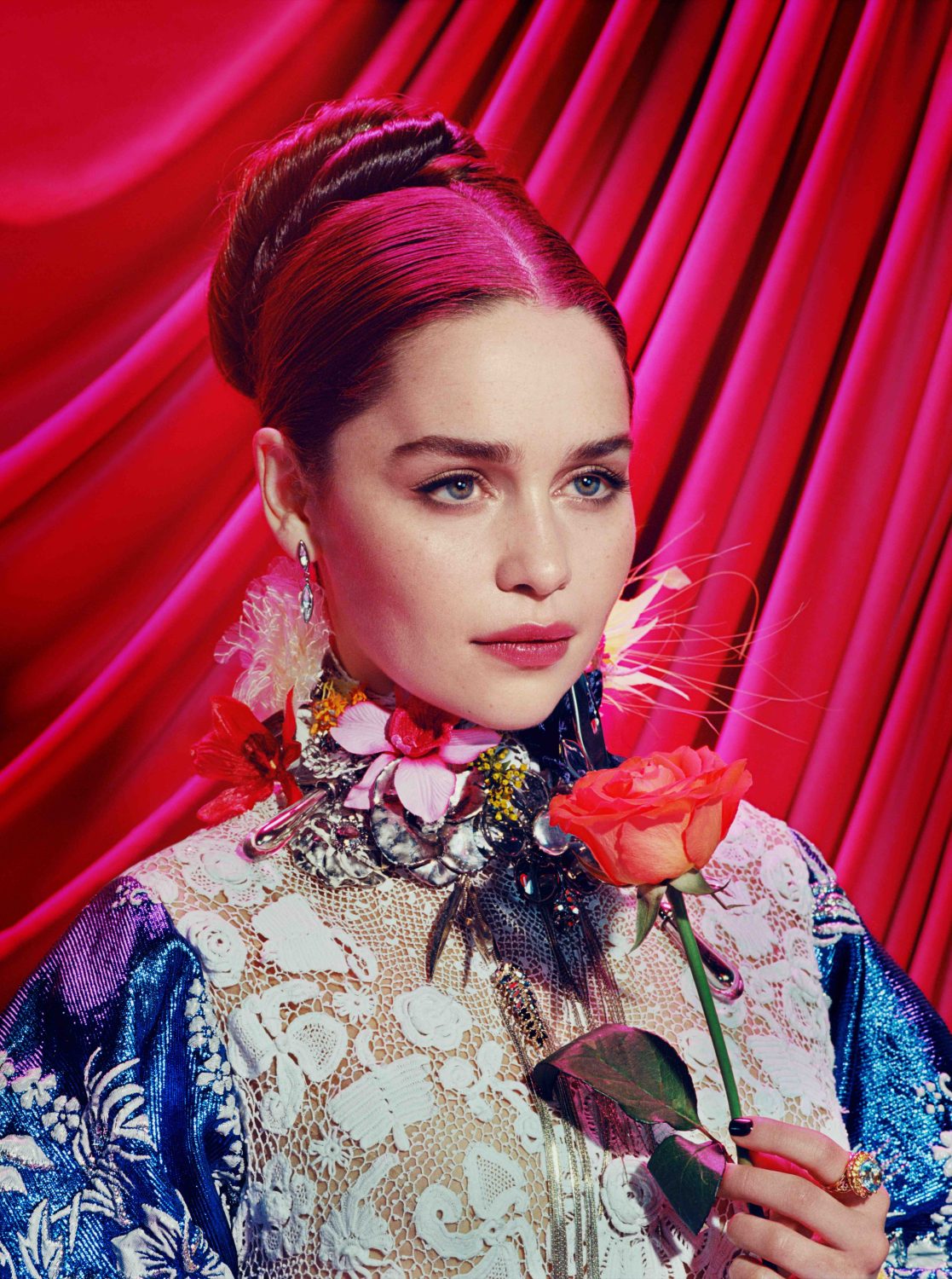 A woman in a blue dress and high heels poses against a pink backdrop in a vibrant studio setting, showcasing bold fashion and makeup.