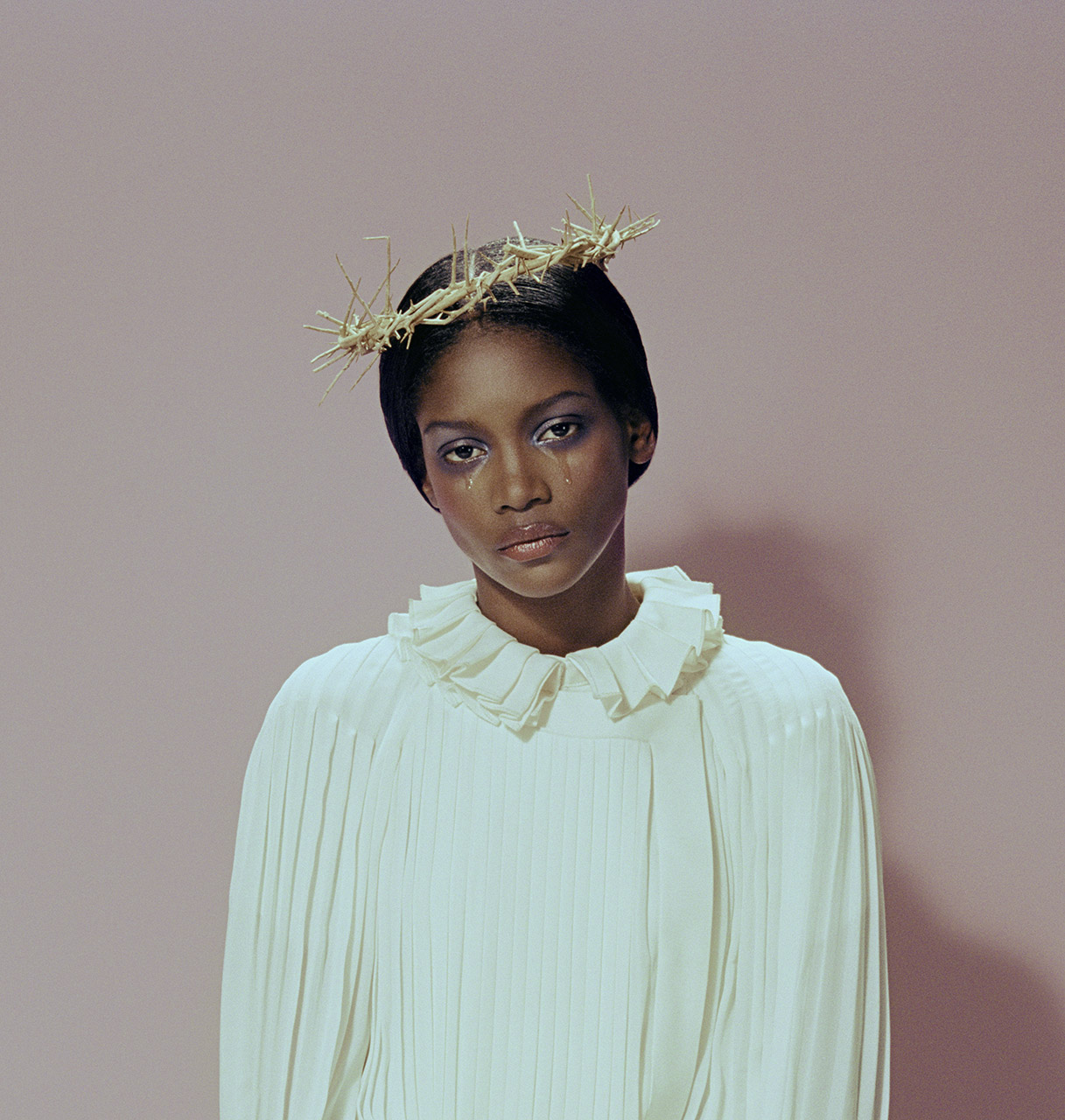 Woman with tear-stained face wearing a crown of thorns and a ruffled collar against a pink background in a portrait shot.