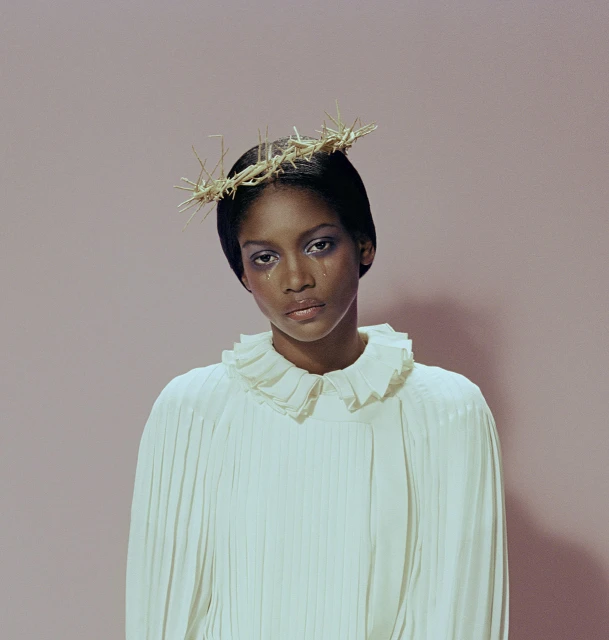Woman with tear-stained face wearing a crown of thorns and a ruffled collar against a pink background in a portrait shot.