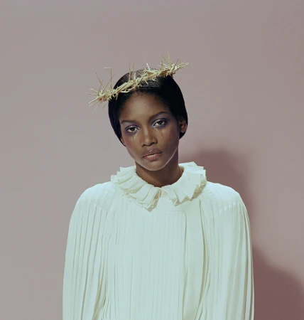 Woman with tear-stained face wearing a crown of thorns and a ruffled collar against a pink background in a portrait shot.