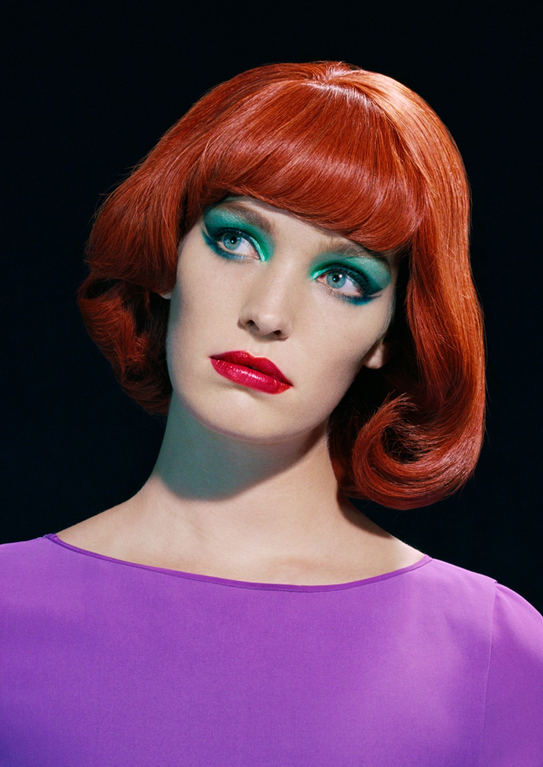 Dramatic fashion photography featuring a woman in a purple dress, a dark hat, and pearls, posing in a modern kitchen with vibrant colors.
