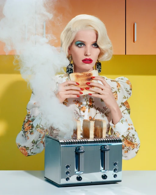 Blonde woman in floral blouse holds burnt toast near toaster in kitchen; cabinets are yellow and orange; smoke around; fashion beauty.