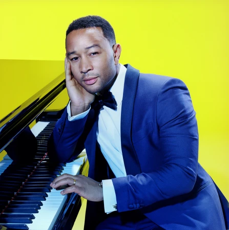 John Legend in a blue tuxedo resting his chin on his hand while seated at a grand piano against a vibrant yellow background