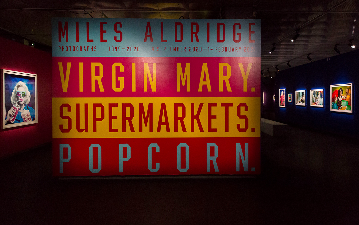 Artwork in gallery featuring a vibrant sign with text and art pieces on the wall, illuminated by gallery lighting in a museum space.