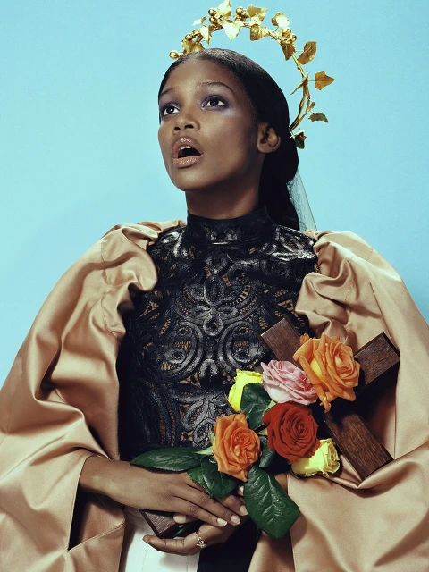 Elegant woman in a black lace top with floral crown and holding a cross and roses, posing against a light blue background.