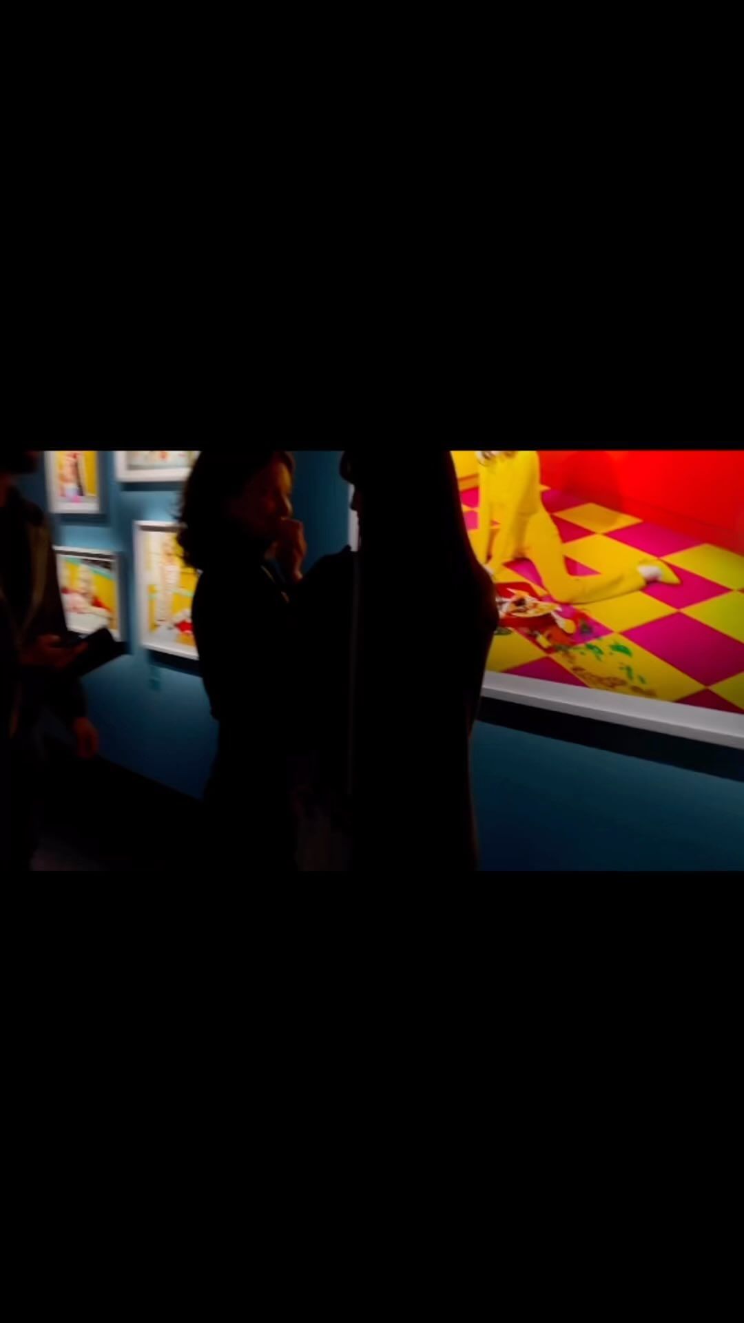 Two silhouetted figures view artwork hanging on a blue wall in a brightly illuminated gallery space with a pink and yellow floor.