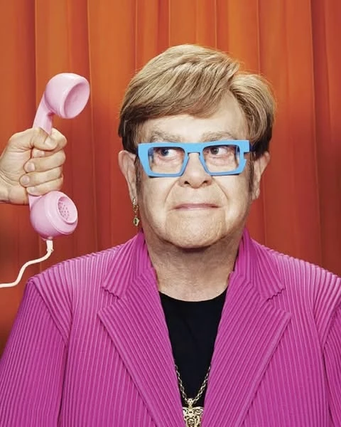 Elderly adult with blonde hair, blue glasses, and a pink jacket poses with a pink telephone against an orange curtain backdrop.