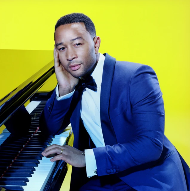 An adult man in a blue tuxedo and bowtie is seated at a black grand piano against a yellow background in a studio portrait.