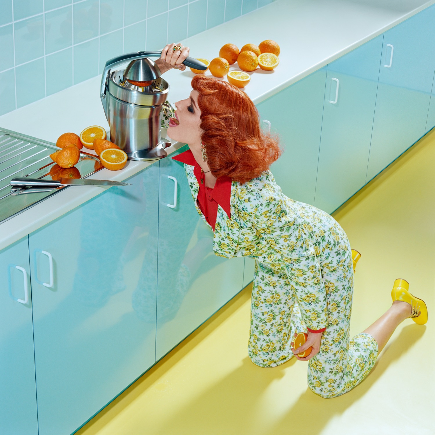 Woman in floral jumpsuit kneeling in a kitchen with a citrus juicer and oranges, vibrant colors, and studio lighting. Food and beverage.