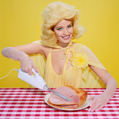 A woman in a yellow dress and blonde wig carves a ham with an electric knife at a red checkered table against a yellow backdrop.