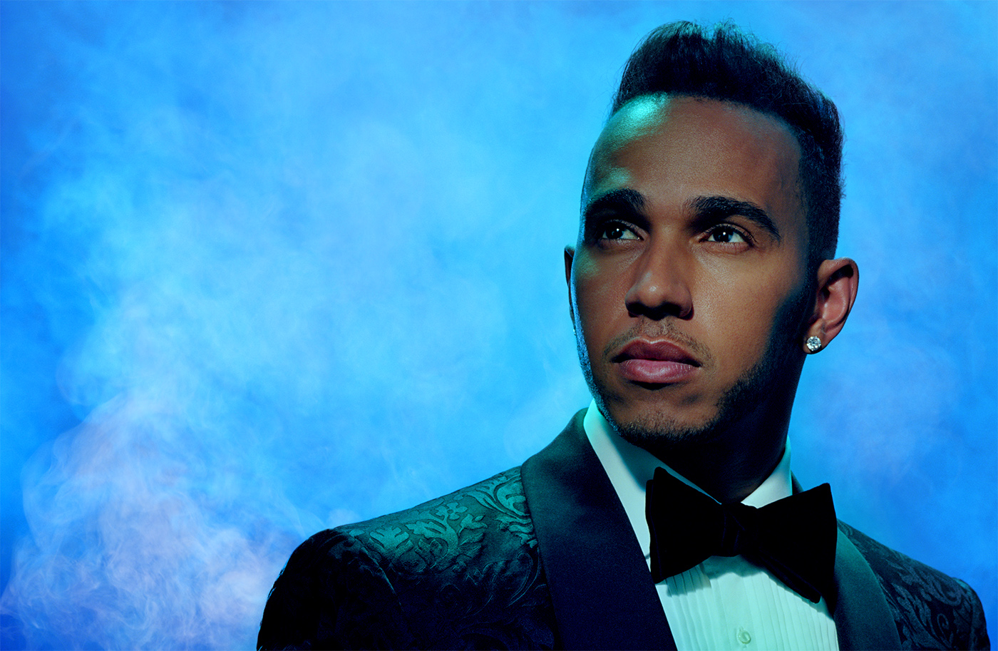Portrait of Lewis Hamilton, wearing a patterned tuxedo and bow tie, against a blue, smoky studio backdrop with soft, diffused lighting.
