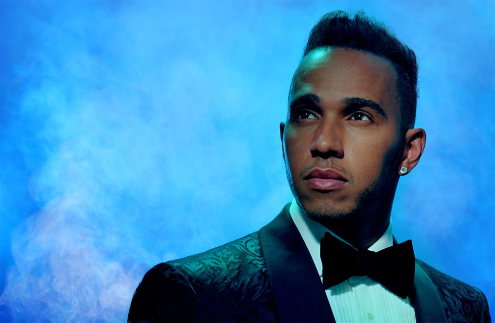 Portrait of Lewis Hamilton, wearing a patterned tuxedo and bow tie, against a blue, smoky studio backdrop with soft, diffused lighting.