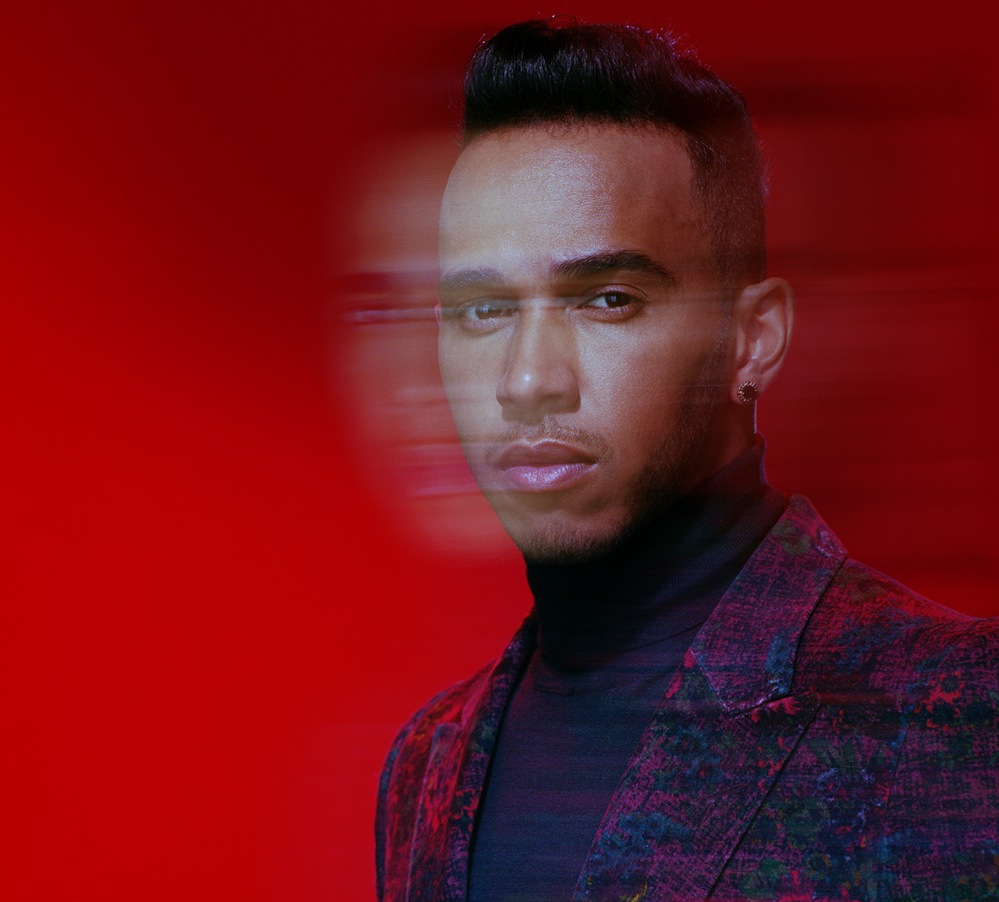 Close-up portrait of an adult male with dark hair wearing a patterned blazer against a solid red background with motion blur effect.