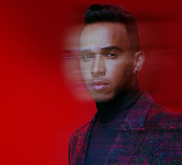 Close-up portrait of an adult male with dark hair wearing a patterned blazer against a solid red background with motion blur effect.