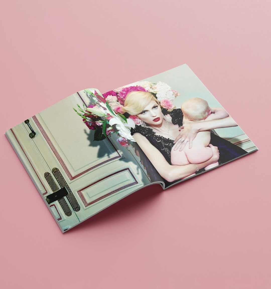 Magazine spread showing a model in an ornate dress, adorned with flowers, holding a baby against a pink background in a studio.