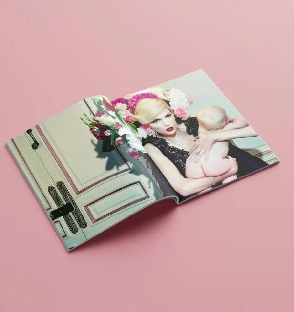 Magazine spread showing a model in an ornate dress, adorned with flowers, holding a baby against a pink background in a studio.